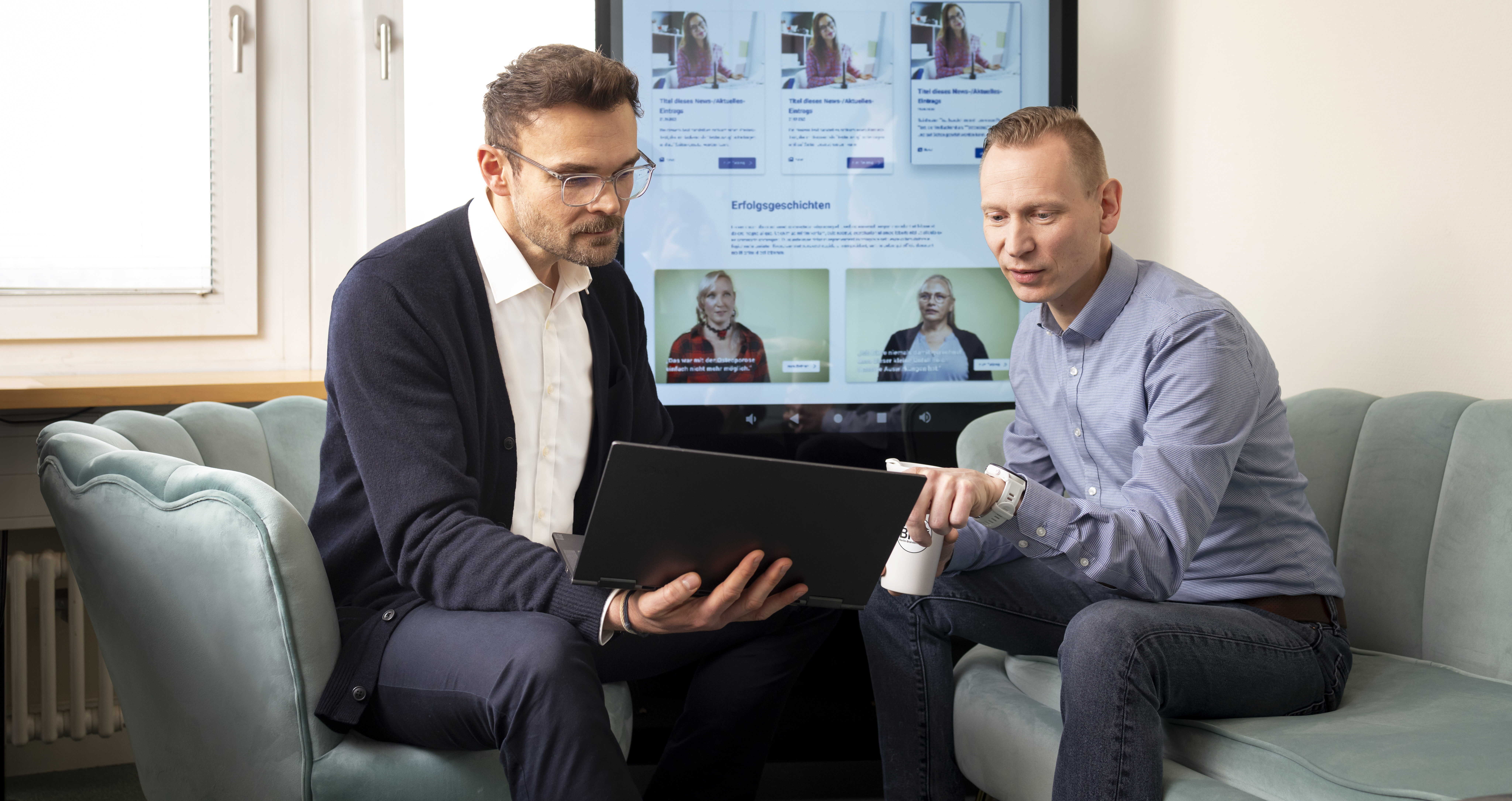 Zwei Personen aus dem Medien- und IT-Umfeld die sich im Büro etwas auf einem Laptop anschauen. Im Hintergrund sieht man eine digitale Werbesäule.